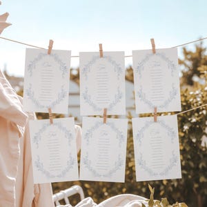 Könnte beinhalten: Sechs weiße Karten mit floralen Designs sind mit Holzklammern an einer Schnur befestigt. Jede Karte hat einen blauen Blumenkranz und Text. Die Karten hängen draußen, mit beigem Stoff und Grün im Hintergrund.