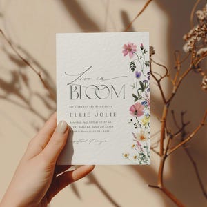May include: A floral invitation card with the words "Love in Bloom" in elegant script. The card features a watercolor floral border with pink, purple, and yellow flowers. The invitation is for a bridal shower.