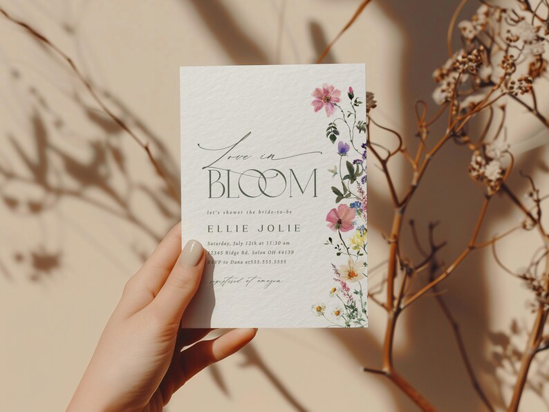 May include: A floral invitation card with the words "Love in Bloom" in elegant script. The card features a watercolor floral border with pink, purple, and yellow flowers. The invitation is for a bridal shower.