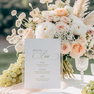 May include: A clear acrylic bar menu with the text "FROM THE Bar" in elegant script. The menu lists wine, liquor, and beer options. A bouquet of white and peach flowers and a bunch of green grapes are in the background.