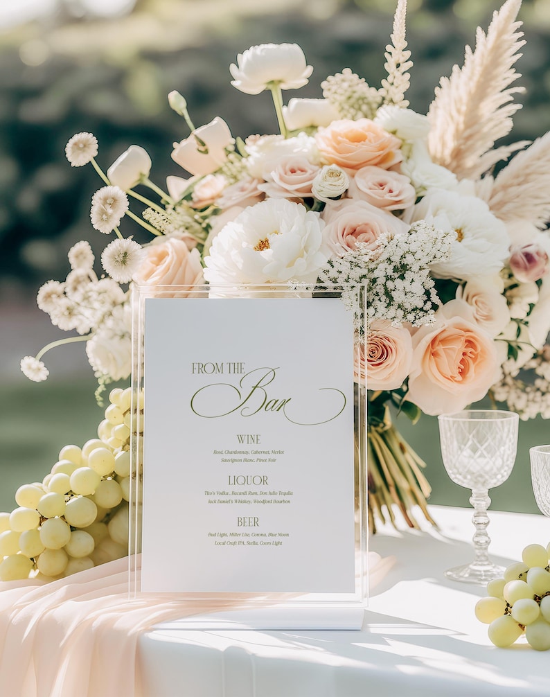 May include: A clear acrylic bar menu with the text "FROM THE Bar" in elegant script. The menu lists wine, liquor, and beer options. A bouquet of white and peach flowers and a bunch of green grapes are in the background.