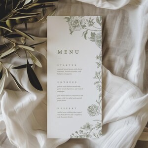 May include: A wedding menu card with a floral design in soft green. The word "MENU" is at the top, with a list of courses below: Starter, Entrees, and Dessert. The card is on a white cloth with olive branches.