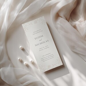 Może przedstawiać: Prostokątna karta z programem ślubnym w kolorze kremowym z ciemnoszarym tekstem szeryfowym o treści Welcome to the wedding of Eloise and Nicholas, April 27, 2024, Naples, Florida oraz The Ceremony. Karta zawiera delikatne kwiatowe wzory i cztery perły.