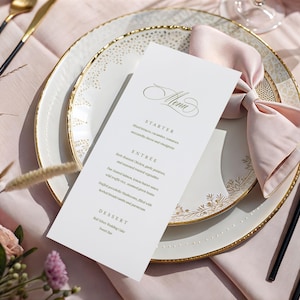 Puede incluir: Elegante mesa con un plato blanco con borde dorado y diseño floral. Una tarjeta de menú con letras doradas descansa sobre el plato, junto con una servilleta rosa y cubiertos dorados con mangos negros. El conjunto está sobre un mantel rosa.