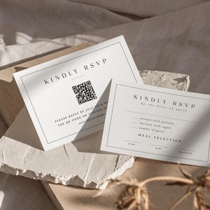 Pu&ograve; includere: Due biglietti RSVP bianchi con testo nero. Un biglietto recita "KINDLY RSVP" e include un codice QR e le istruzioni per la risposta. L'altro biglietto presenta i dettagli RSVP e le opzioni di selezione dei pasti. I biglietti sono su una superficie neutra.
