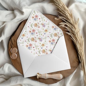 May include: A white envelope with a floral interior, showcasing pink, blue, and yellow flowers on a white backdrop. The envelope is open, displaying the design. Two gold wax seals and a wooden stamp are also present.