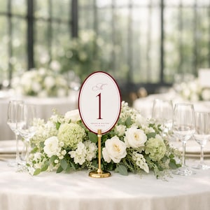 Puede incluir: Decoraci&oacute;n de mesa de boda con centro floral y n&uacute;mero de mesa. El n&uacute;mero de mesa ovalado es blanco con borde burdeos y el n&uacute;mero "1" en burdeos. El centro de mesa incluye rosas blancas y vegetaci&oacute;n.