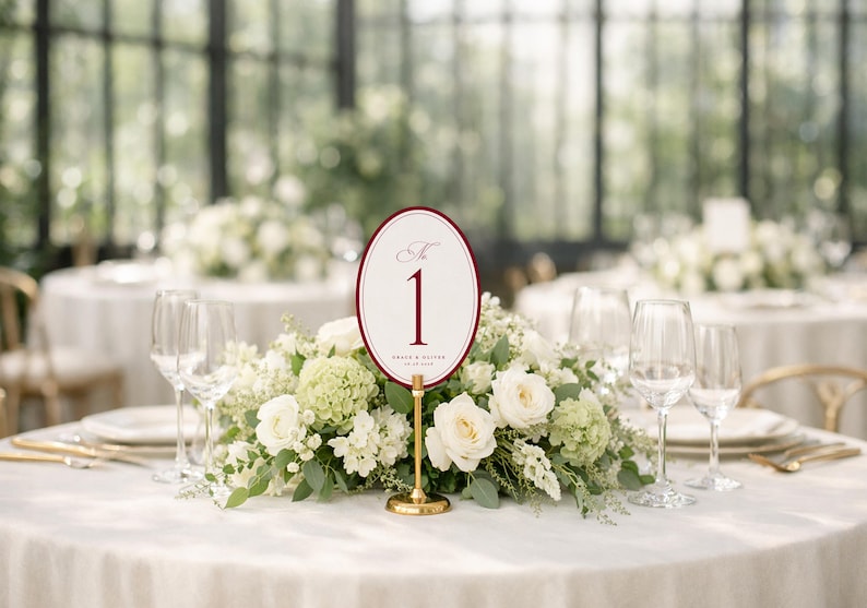 Puede incluir: Decoraci&oacute;n de mesa de boda con centro floral y n&uacute;mero de mesa. El n&uacute;mero de mesa ovalado es blanco con borde burdeos y el n&uacute;mero "1" en burdeos. El centro de mesa incluye rosas blancas y vegetaci&oacute;n.