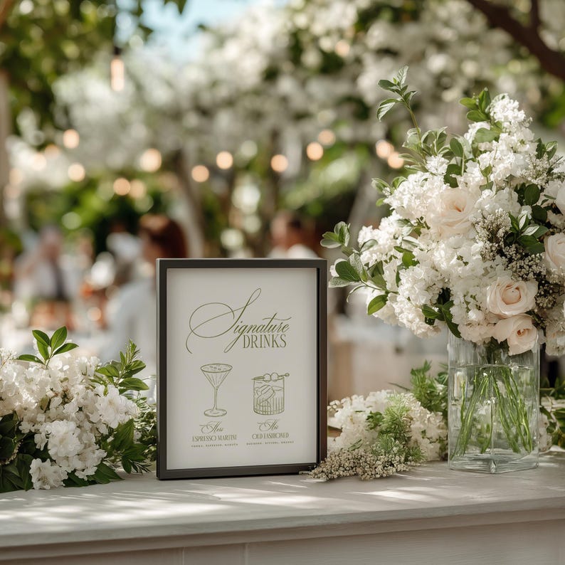 Puede incluir: Un letrero con marco negro con las palabras "Signature Drinks" e ilustraciones de c&oacute;cteles. El letrero est&aacute; sobre una superficie blanca, rodeado de flores blancas y un jarr&oacute;n de rosas blancas. El fondo es borroso con vegetaci&oacute;n.