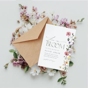 May include: A white invitation card with the words "Love in Bloom" and floral accents, accompanied by a brown envelope and surrounded by pink and white flowers. The invitation includes event details.