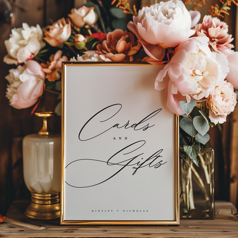 May include: A white sign with a gold frame and black calligraphy that reads "Cards and Gifts" with the names "Kinsley + Nicholas" at the bottom. The sign is surrounded by pink and peach flowers.
