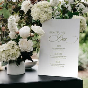 May include: A white bar menu with the text "FROM THE Bar" in elegant script, listing wine, liquor, and beer options. The menu is set against a backdrop of white flowers in a white vase, creating a sophisticated ambiance.