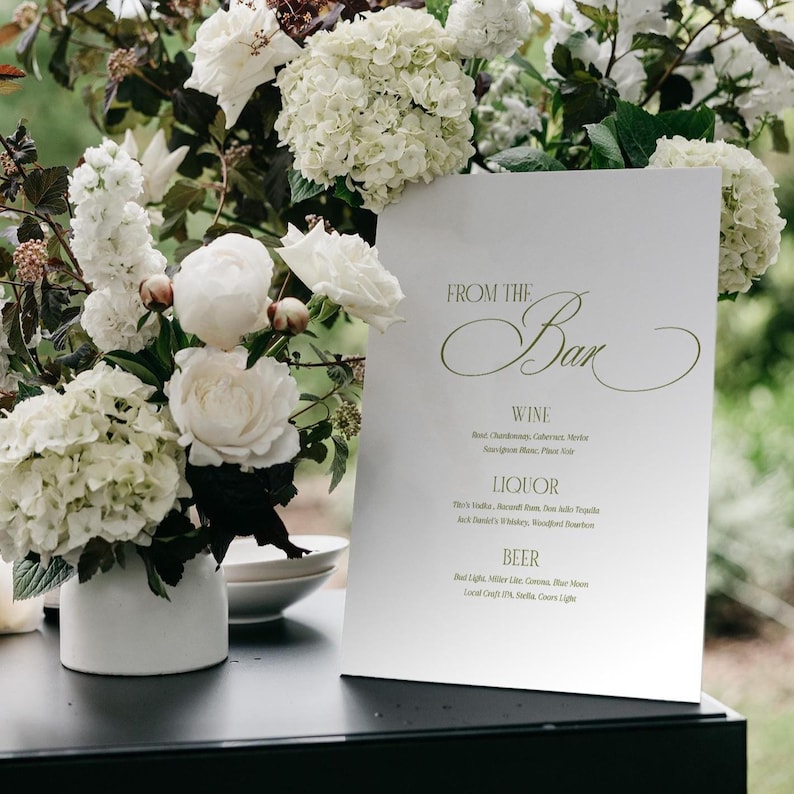 May include: A white bar menu with the text "FROM THE Bar" in elegant script, listing wine, liquor, and beer options. The menu is set against a backdrop of white flowers in a white vase, creating a sophisticated ambiance.
