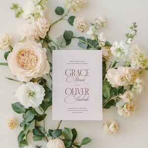Puede incluir: Una invitaci&oacute;n de boda con los nombres "Grace Atwood" y "Oliver Sembriche" en escritura burdeos, rodeada de flores crema y blancas y follaje verde. La invitaci&oacute;n incluye la fecha y el lugar de la boda.