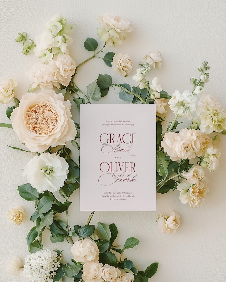 Puede incluir: Una invitaci&oacute;n de boda con los nombres "Grace Atwood" y "Oliver Sembriche" en escritura burdeos, rodeada de flores crema y blancas y follaje verde. La invitaci&oacute;n incluye la fecha y el lugar de la boda.