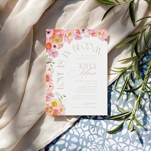 May include: A wedding invitation with floral accents. The invitation features the text "Love is in Bloom" and the name "Kayla Morrison". The invitation is decorated with pink, yellow, and white flowers. The background is a light beige fabric and a blue floral pattern.
