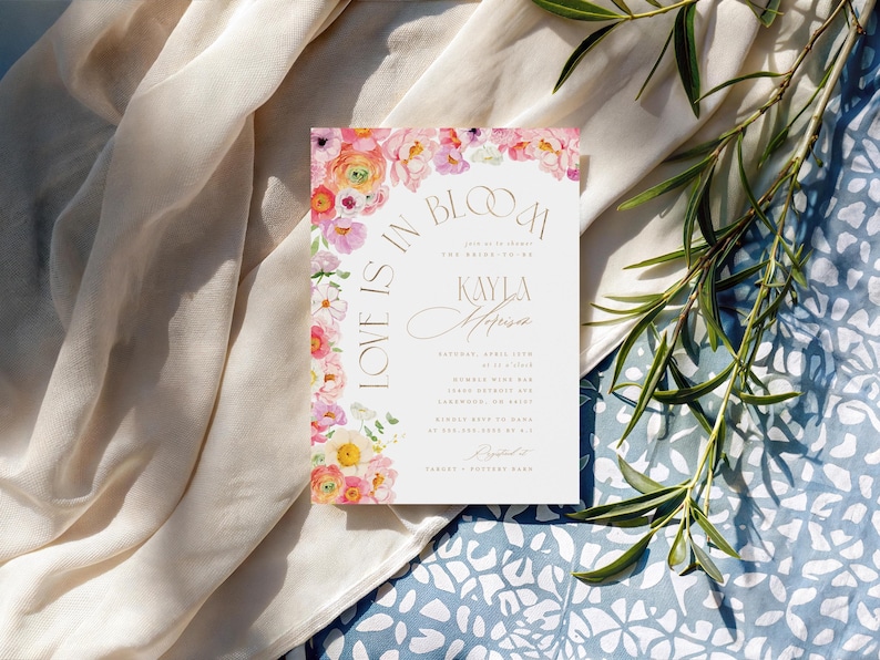 May include: A wedding invitation with floral accents. The invitation features the text "Love is in Bloom" and the name "Kayla Morrison". The invitation is decorated with pink, yellow, and white flowers. The background is a light beige fabric and a blue floral pattern.