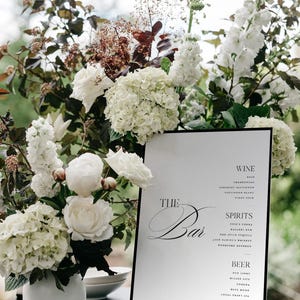 May include: A white bar menu sign with black text reading The Bar, Wine, Spirits, and Beer, displayed next to a white ceramic vase filled with white hydrangeas, white roses, and dark foliage. The sign lists various alcoholic beverage options.