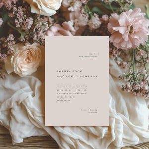 May include: A wedding invitation with a beige background and brown text. The invitation reads "Sophia Sogo and Luke Thompson" with the wedding date and location below. The invitation is surrounded by pink and white flowers.