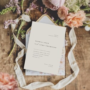 May include: A wedding invitation card with a cream background and black text. The invitation reads "Sophia Sogo and Luke Thompson" and includes the wedding date, time, and location. The card is surrounded by pink and white flowers and a white ribbon.