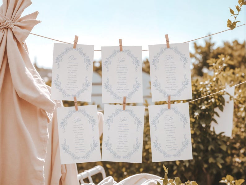 May include: Six white rectangular cards with blue floral borders and printed text, each suspended by a small wooden clothespin from a horizontal string. The cards are arranged in two rows of three, displaying lists of names within the floral frames.