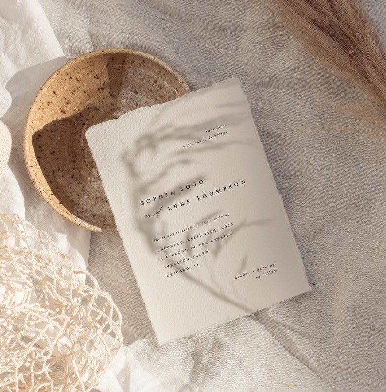 May include: A minimalist wedding invitation with a cream background and black text. The invitation reads "Sophia Sogo and Luke Thompson" and includes the wedding date, time, and location. The invitation is on a textured surface with a bowl and dried grass.