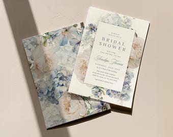 May include: Bridal shower invitation set with a floral design of white and blue hydrangeas and roses. The invitation card includes the text "Bridal Shower" and event details. The set is on a light beige surface.