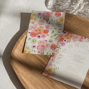 May include: Two floral wedding invitations with watercolour flowers in pink, yellow, and white. One invitation has the text "LOVE IS IN BLOOM" and the name "Kayla" in gold script. The invitations are on a wooden tray.