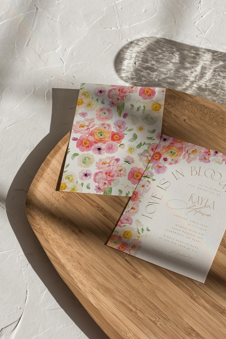 May include: Two floral wedding invitations with watercolour flowers in pink, yellow, and white. One invitation has the text "LOVE IS IN BLOOM" and the name "Kayla" in gold script. The invitations are on a wooden tray.