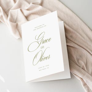 Puede incluir: Un programa de boda blanco plegado con una elegante escritura que dice "Grace and Oliver". El programa incluye la fecha y el lugar de la boda en una delicada fuente verde oliva. Un tejido beige est&aacute; drapeado en el fondo.