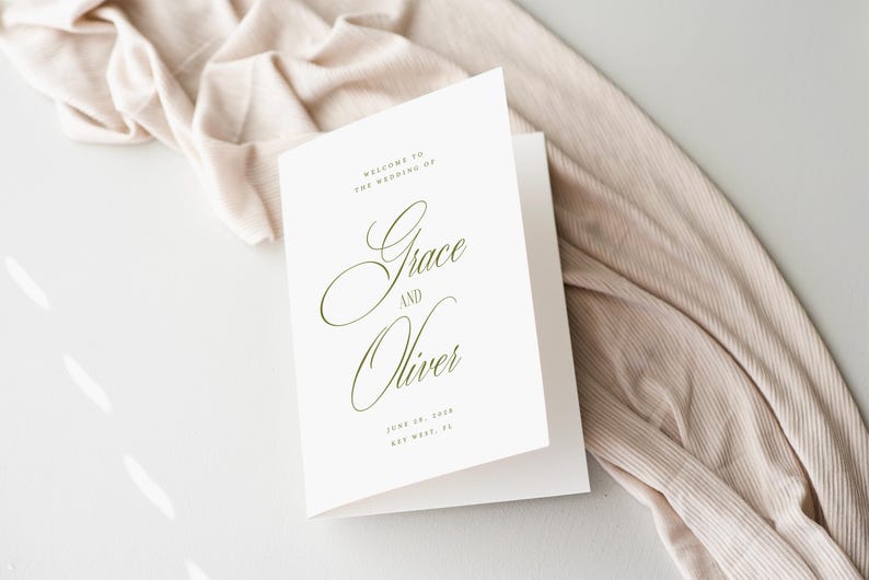 Puede incluir: Un programa de boda blanco plegado con una elegante escritura que dice "Grace and Oliver". El programa incluye la fecha y el lugar de la boda en una delicada fuente verde oliva. Un tejido beige est&aacute; drapeado en el fondo.