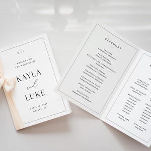 Op de afbeelding: Trouwceremonieprogramma's met zwarte tekst op wit papier. E&eacute;n programma is gevouwen en vastgebonden met een cr&egrave;mekleurig lint. De tekst bevat de namen Kayla en Luke en de datum 22 april 2028.