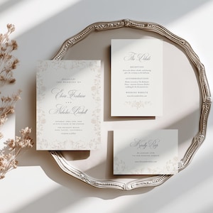 May include: A set of three white wedding stationery cards with elegant black serif and script typography, featuring a faint floral border design. The cards include an invitation, a details card, and an RSVP card, all displayed on a silver tray.