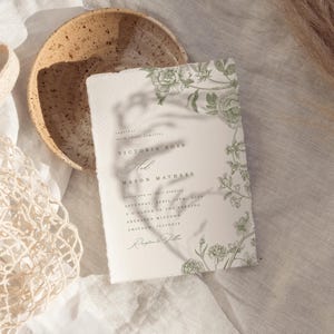 May include: A rectangular wedding invitation card with deckled edges, featuring green botanical illustrations of flowers and leaves. The text reads: Together with their families Victoria Ross and Mason Mathers invite you to their wedding.
