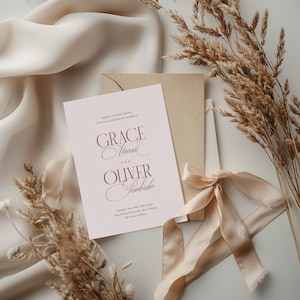 Puede incluir: Una invitaci&oacute;n de boda con los nombres "Grace Atwood" y "Oliver Sembriche" en escritura burdeos. La invitaci&oacute;n est&aacute; en una tarjeta rosa claro, con un sobre y una cinta beige. Espigas de trigo secas y una tela beige en el fondo.