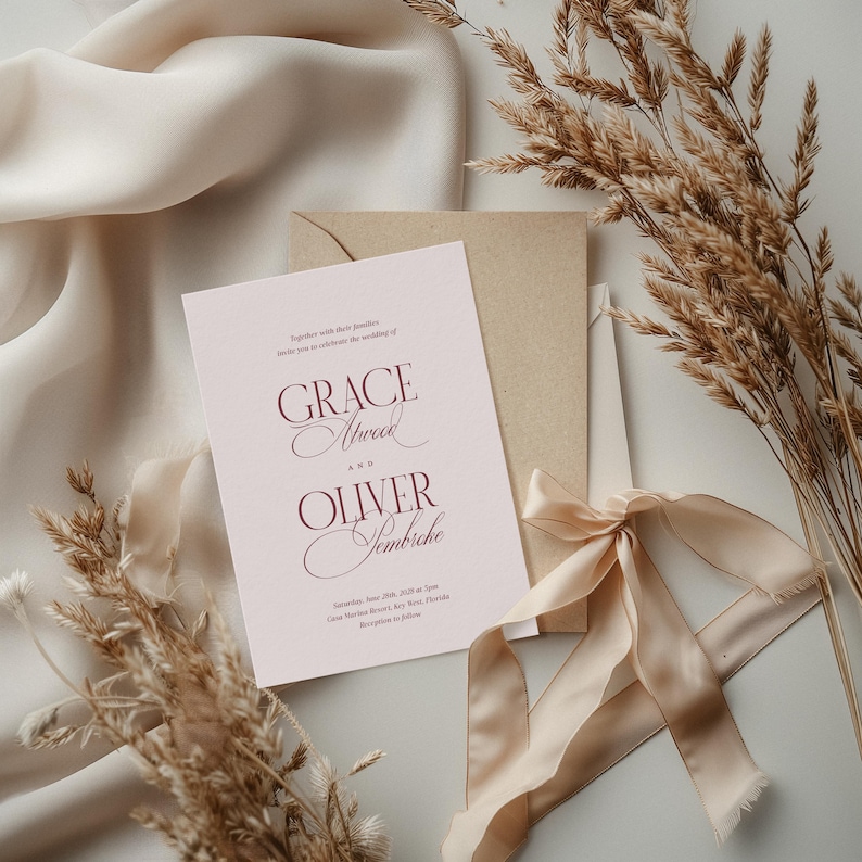 Puede incluir: Una invitaci&oacute;n de boda con los nombres "Grace Atwood" y "Oliver Sembriche" en escritura burdeos. La invitaci&oacute;n est&aacute; en una tarjeta rosa claro, con un sobre y una cinta beige. Espigas de trigo secas y una tela beige en el fondo.