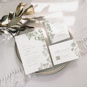 May include: A wedding invitation suite featuring three white cards with sage green botanical illustrations of roses and leaves. The text includes names Victoria Ross and Mason Mathews, event details, and a QR code on the response card.