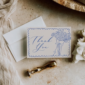 May include: A white thank you card with blue floral illustration and text. The card has a scalloped border and is accompanied by a white envelope and a gold wax seal. A bouquet of flowers is depicted on the card.