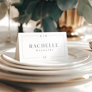May include: A table setting with white plates and gold rims, featuring a name card that reads "RACHELLE Reynolds" and the number "13". The card is on a stack of plates, with a gold spoon visible.