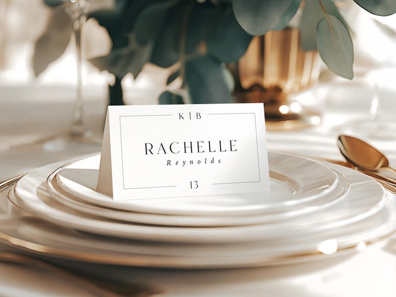 May include: A table setting with white plates and gold rims, featuring a name card that reads "RACHELLE Reynolds" and the number "13". The card is on a stack of plates, with a gold spoon visible.