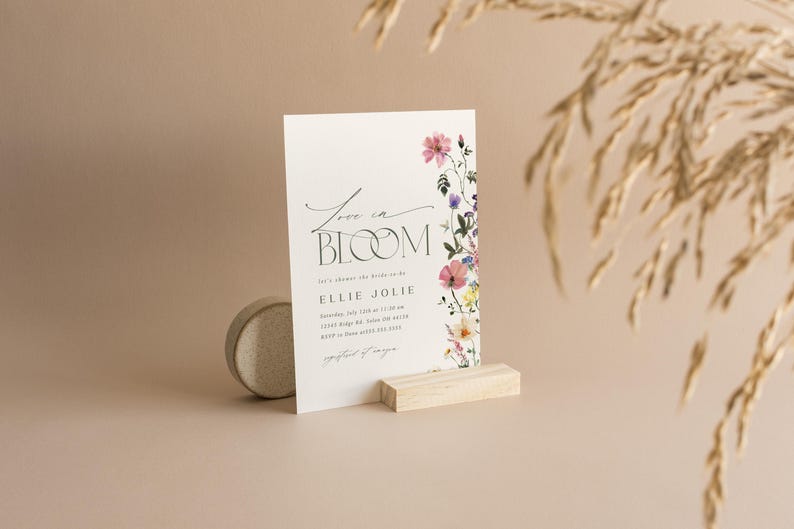 May include: A white invitation card with the text "Love in Bloom" and floral accents. The card is propped up on a wooden block, with a small, round, stone-like object to the left. The background is a soft, neutral color.