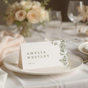 May include: A white place card with the name "AMELIA WESTLEY" printed in green, along with "table one." Floral design in the upper right corner. The card is on a white plate, with a blurred floral arrangement in the background.