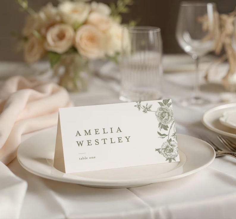 May include: A white place card with the name "AMELIA WESTLEY" printed in green, along with "table one." Floral design in the upper right corner. The card is on a white plate, with a blurred floral arrangement in the background.