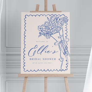 Könnte beinhalten: Ein cremefarbenes Schild auf einer hölzernen Staffelei mit blauer Strichzeichnung. Auf dem Schild steht "welcome to Ellie's Bridal Shower" mit einer floralen Bouquet-Illustration. Das Schild ist mit einem gewellten Rand und dem Text "off the market since 2023" umrandet.
