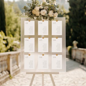 May include: A clear acrylic seating plan with nine numbered cards, each secured by a gold pin. The chart is displayed on a white easel, adorned with a floral arrangement of white roses and foliage. The background is a blurred outdoor scene.