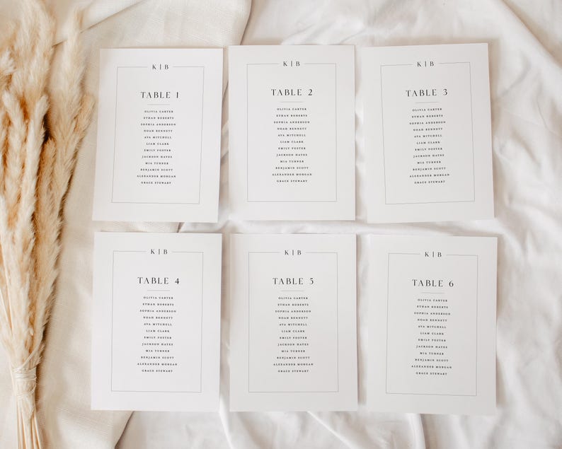 May include: Six white rectangular table cards with black text, arranged on a white surface. Each card is labelled with a table number and a list of names. The cards have a minimalist design, suitable for weddings or events.