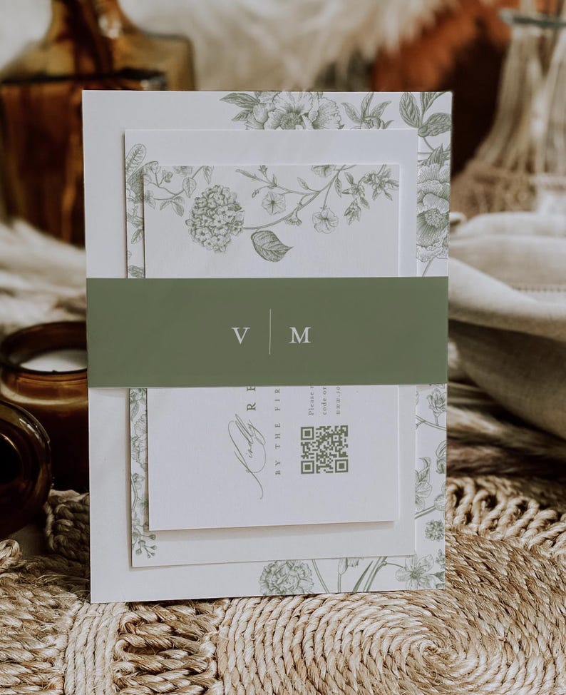 May include: A layered invitation suite featuring a floral design in shades of green and white. The suite includes a belly band with the initials "V M". The invitation has a QR code and text. The overall theme is elegant and botanical.