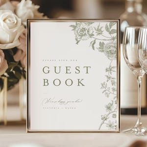 May include: A framed guest book sign with a gold border. The sign has the words "GUEST BOOK" in a serif font. The sign is decorated with floral illustrations in a muted green color. The text "We're so happy you're here!" is written below.
