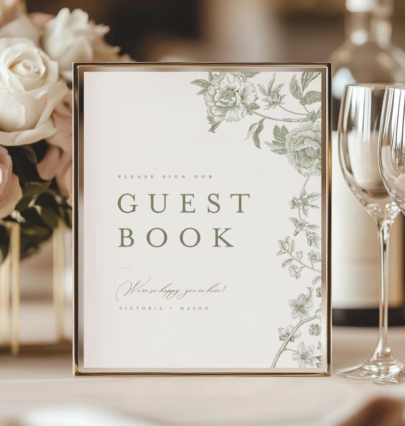 May include: A framed guest book sign with a gold border. The sign has the words "GUEST BOOK" in a serif font. The sign is decorated with floral illustrations in a muted green color. The text "We're so happy you're here!" is written below.