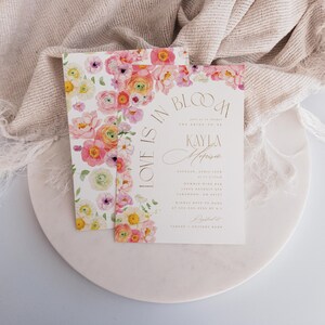 May include: Two wedding invitations with floral designs. The invitations feature a watercolour floral border in shades of pink, yellow, and white. The text on the invitation reads "Love is in Bloom" and includes the names "Kayla Melissa".